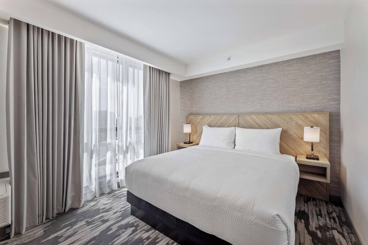 A hotel room with a large bed, two bedside tables with lamps, a wooden headboard, and a large window with curtains.