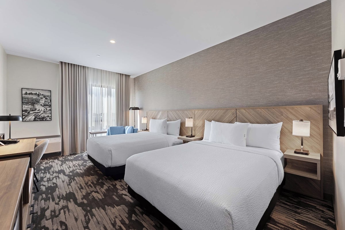 A hotel room with two beds, a desk, and a chair. There are two bedside tables with lamps.