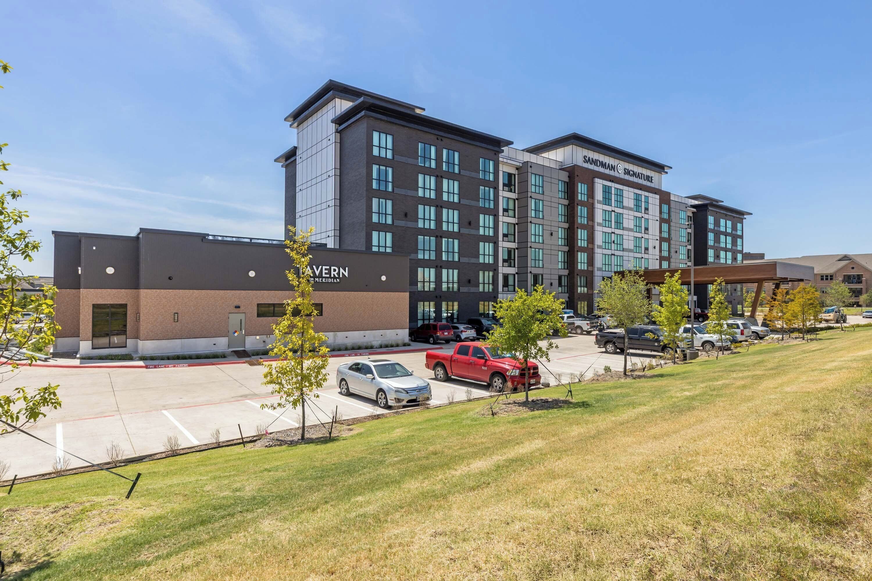 A multi-story hotel with a modern design, featuring a parking lot with several vehicles and a grassy area in the foreground.