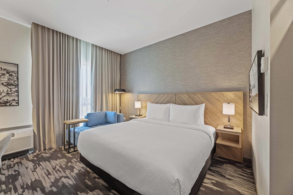 A hotel room with a large bed, a desk with a chair, a lamp, and a wall-mounted TV.
