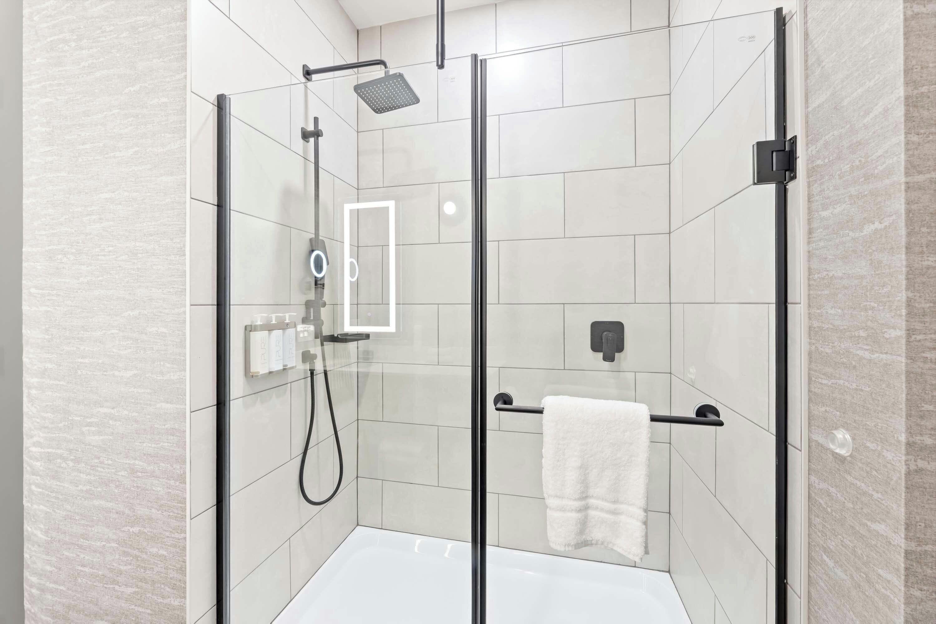 Een moderne douchecel met een glazen deur, een handdouche, een handdoekhouder en een zeepdispenser.
