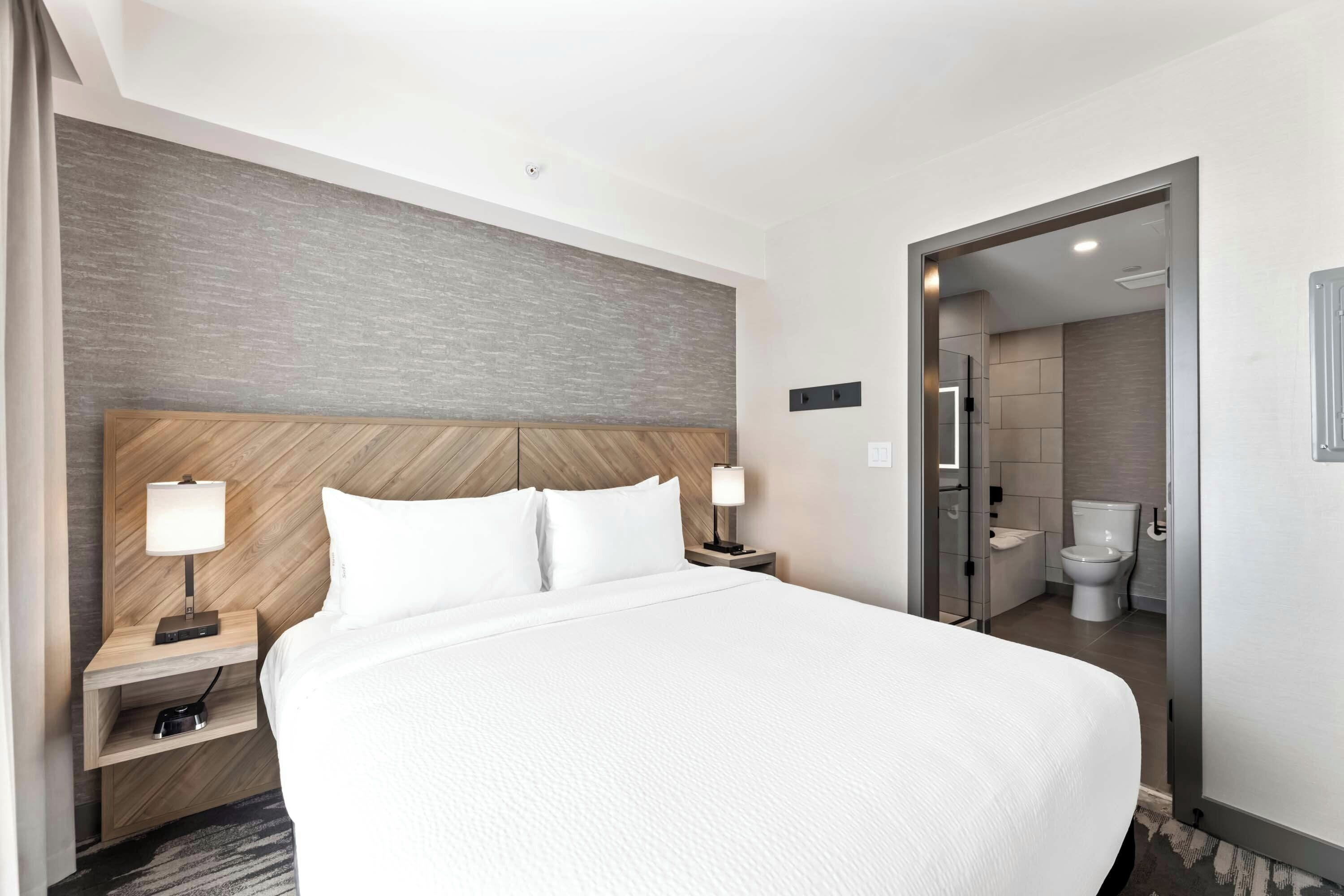 A hotel room with a large bed, bedside tables with lamps, a wooden headboard, and a bathroom visible through an open door.