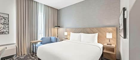 A hotel room with a large bed, a desk, a chair, a nightstand, a lamp, and a wall-mounted air conditioning unit.