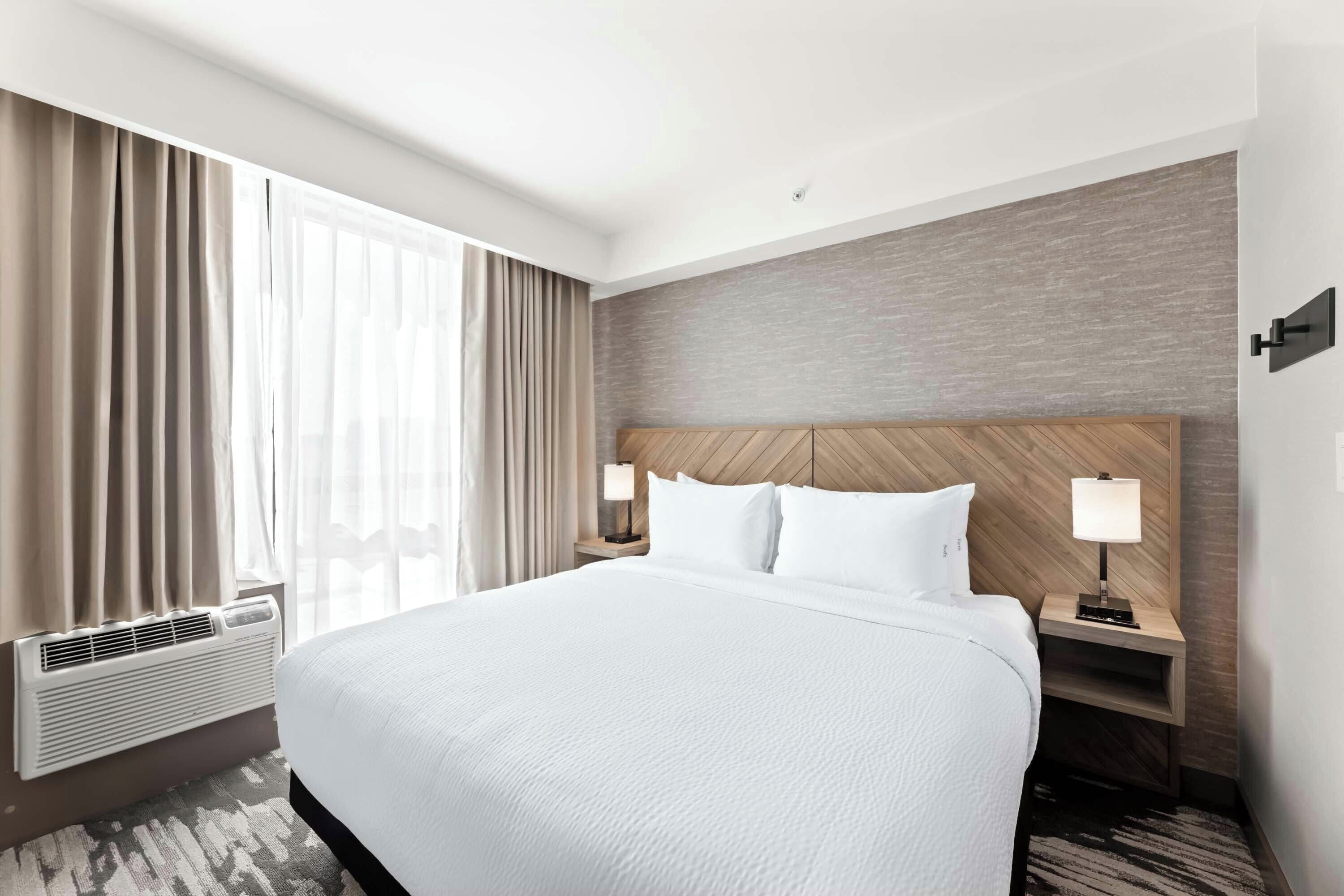 Une chambre d’hôtel avec un grand lit, deux tables de chevet avec des lampes, une fenêtre avec des rideaux et un mur à la finition texturée.