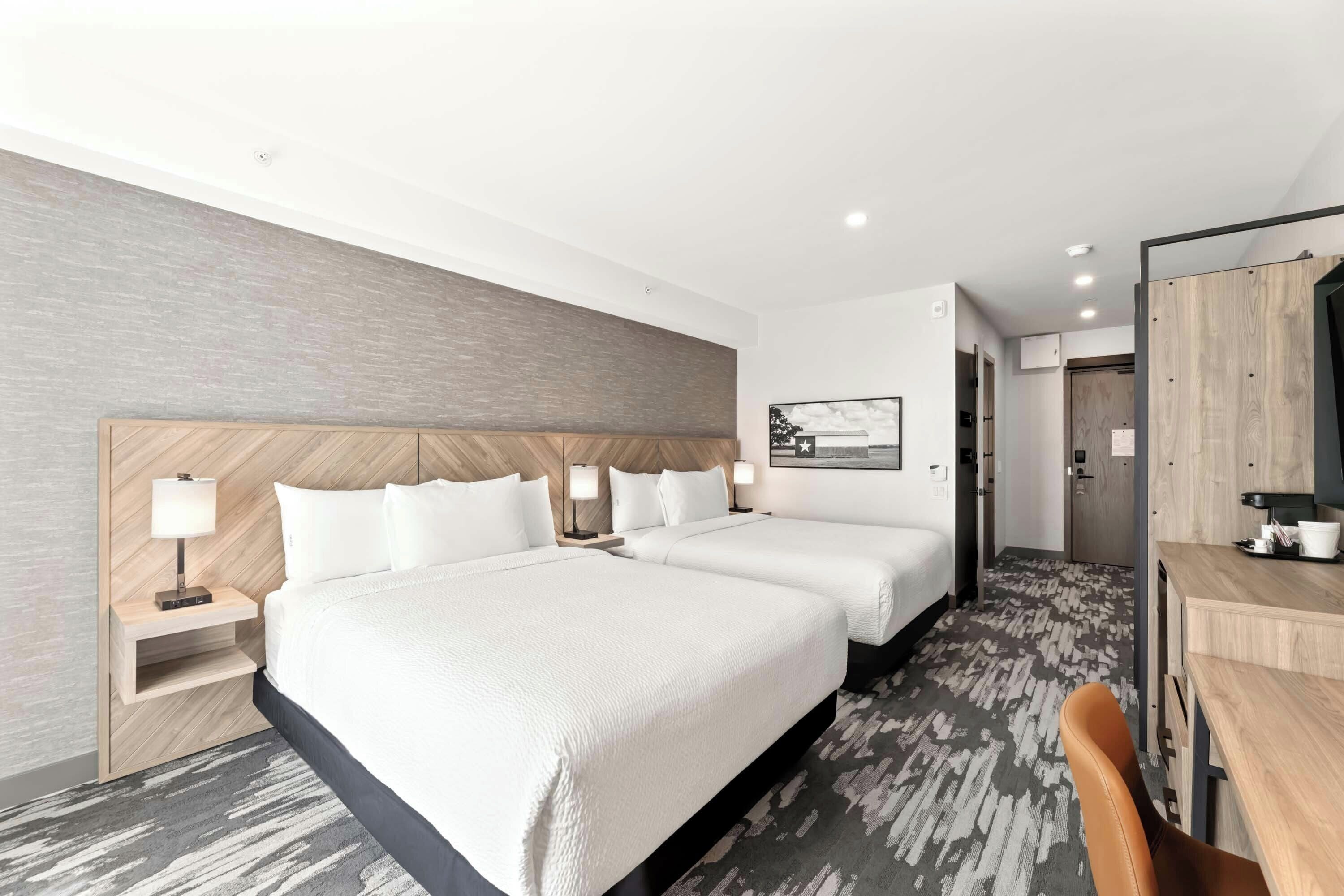 Quarto de hotel com duas camas, uma escrivaninha, uma cadeira, um guarda-roupa e um quadro na parede.
