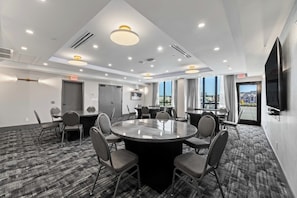 Uma sala de conferências moderna com uma grande mesa redonda, várias cadeiras, luminárias no teto e uma televisão na parede.