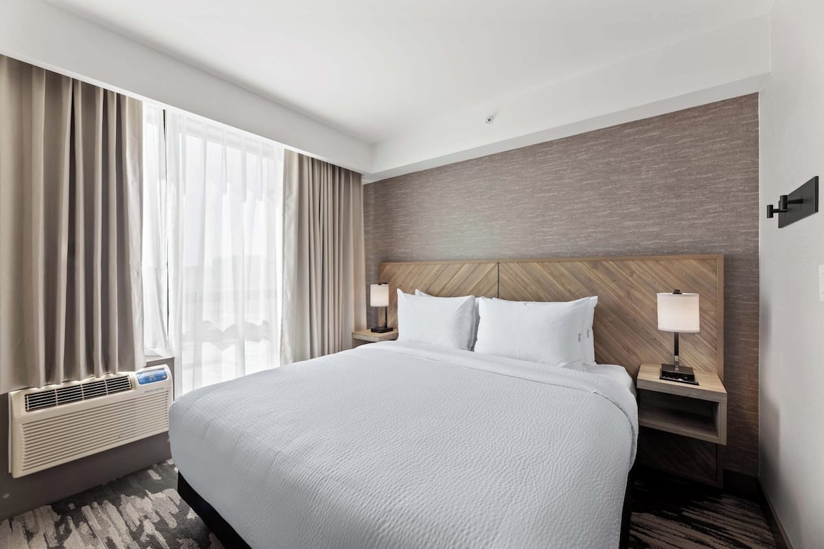 A hotel room with a large bed, two bedside tables with lamps, a wall-mounted heater, and a window with curtains.