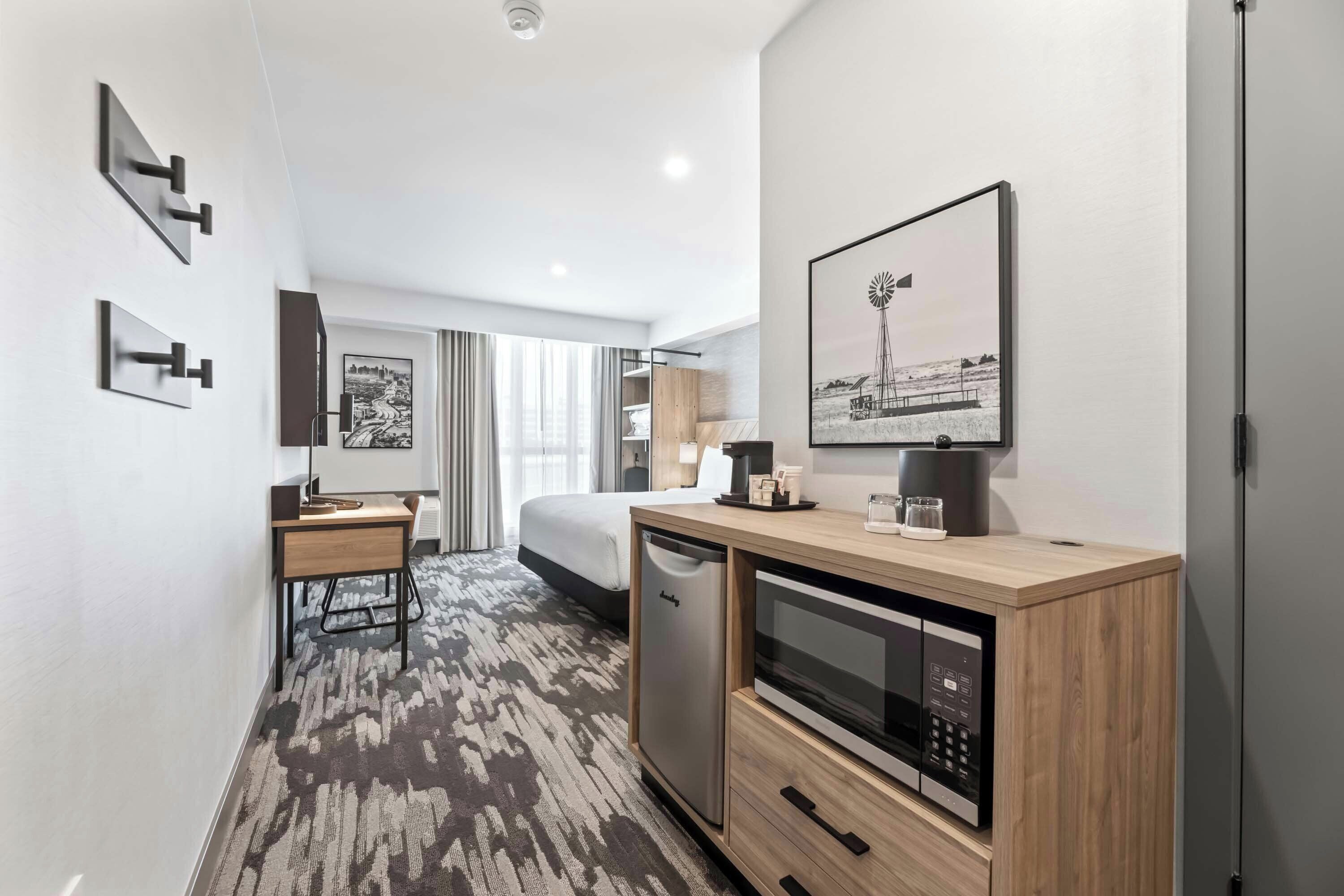 A modern hotel room with a bed, a desk, a microwave, a coffee maker, and a large window with curtains.