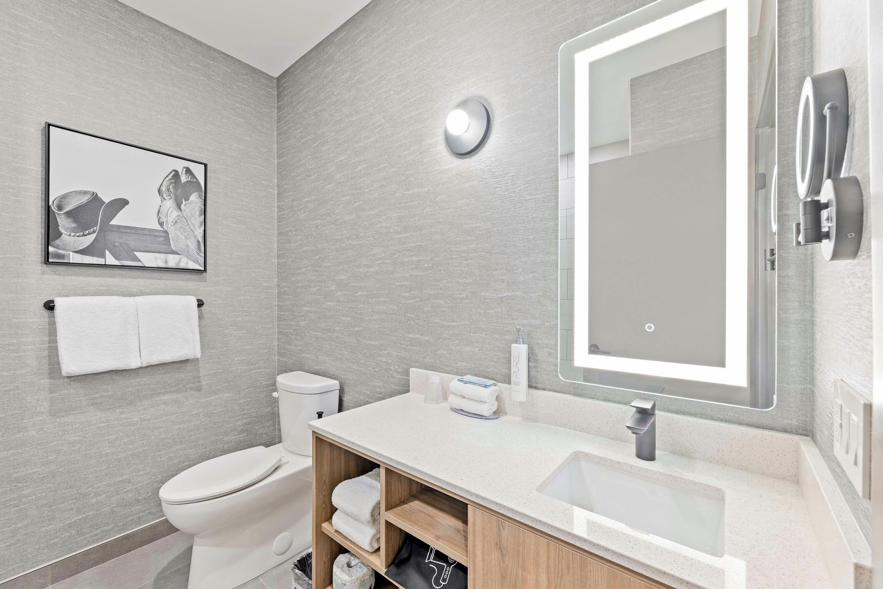 A modern bathroom with a toilet, sink, and mirror. There is a framed picture on the wall and a towel rack.