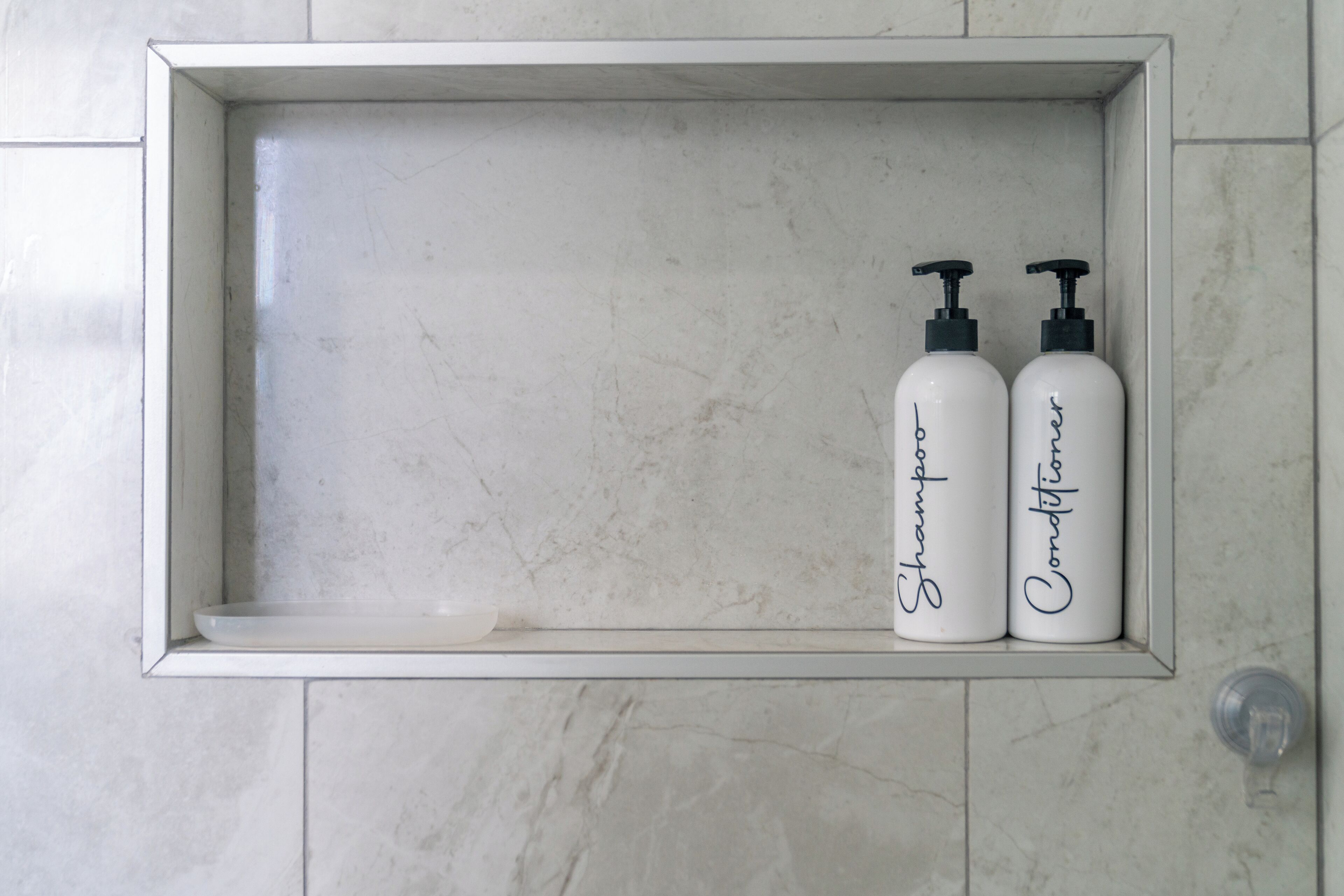 Une étagère de salle de bain avec deux bouteilles blanches étiquetées « Shampoing » et « Après-shampoing », le tout placé devant un mur carrelé.