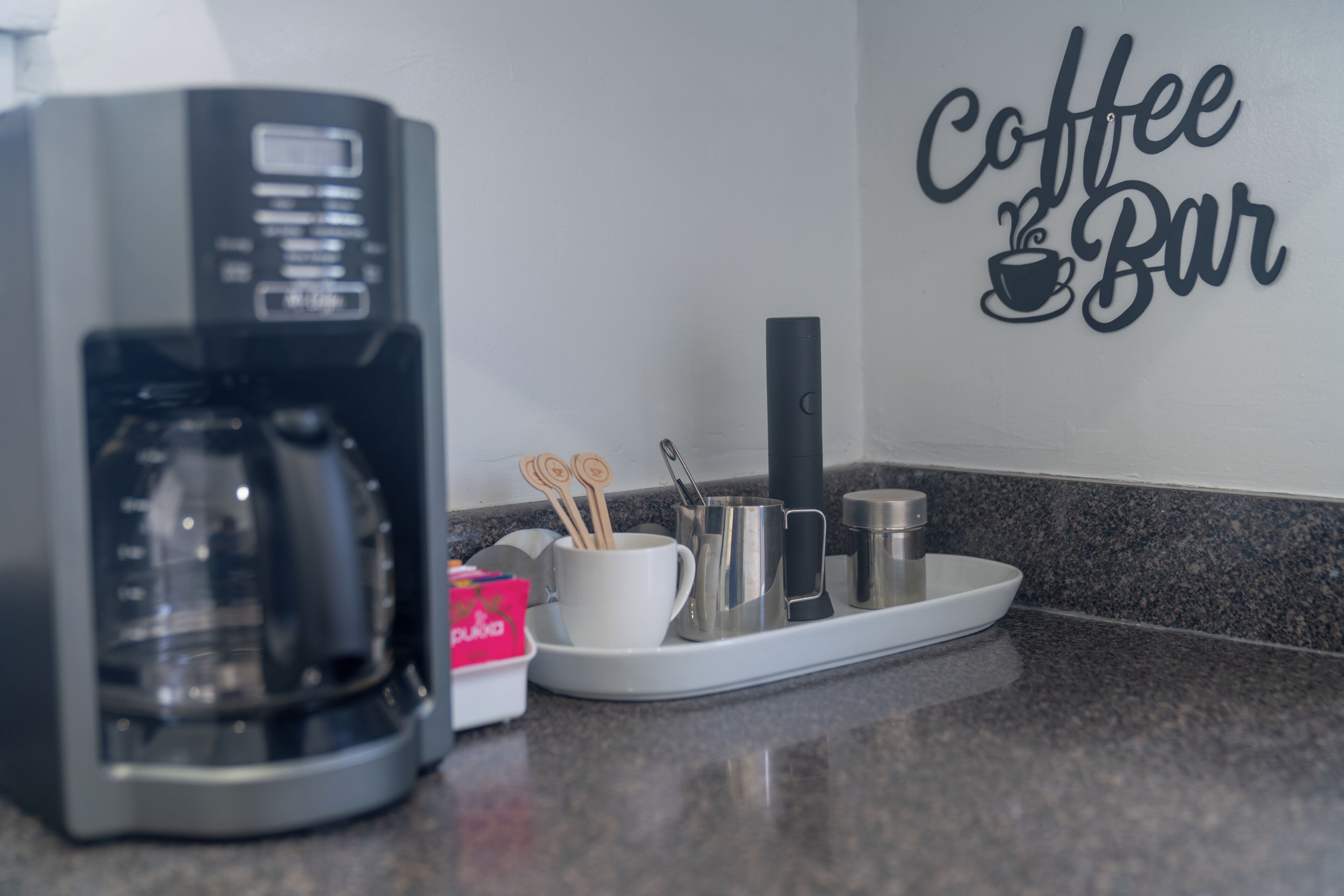 A coffee maker, a tray with utensils, and a wall sign that reads "Coffee Bar".
