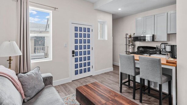 A compact living space with a kitchenette, a dining area with bar stools, a sofa with a throw blanket, and a window with curtains.