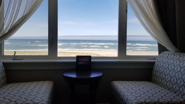 A room with a view of the beach, two patterned chairs, a small table with a sign, and sheer curtains. - Sail Away - Waldport (Waldport)