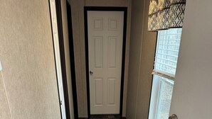 Un couloir doté d’un plancher en bois, d’une porte blanche et d’une fenêtre avec un store à motifs.