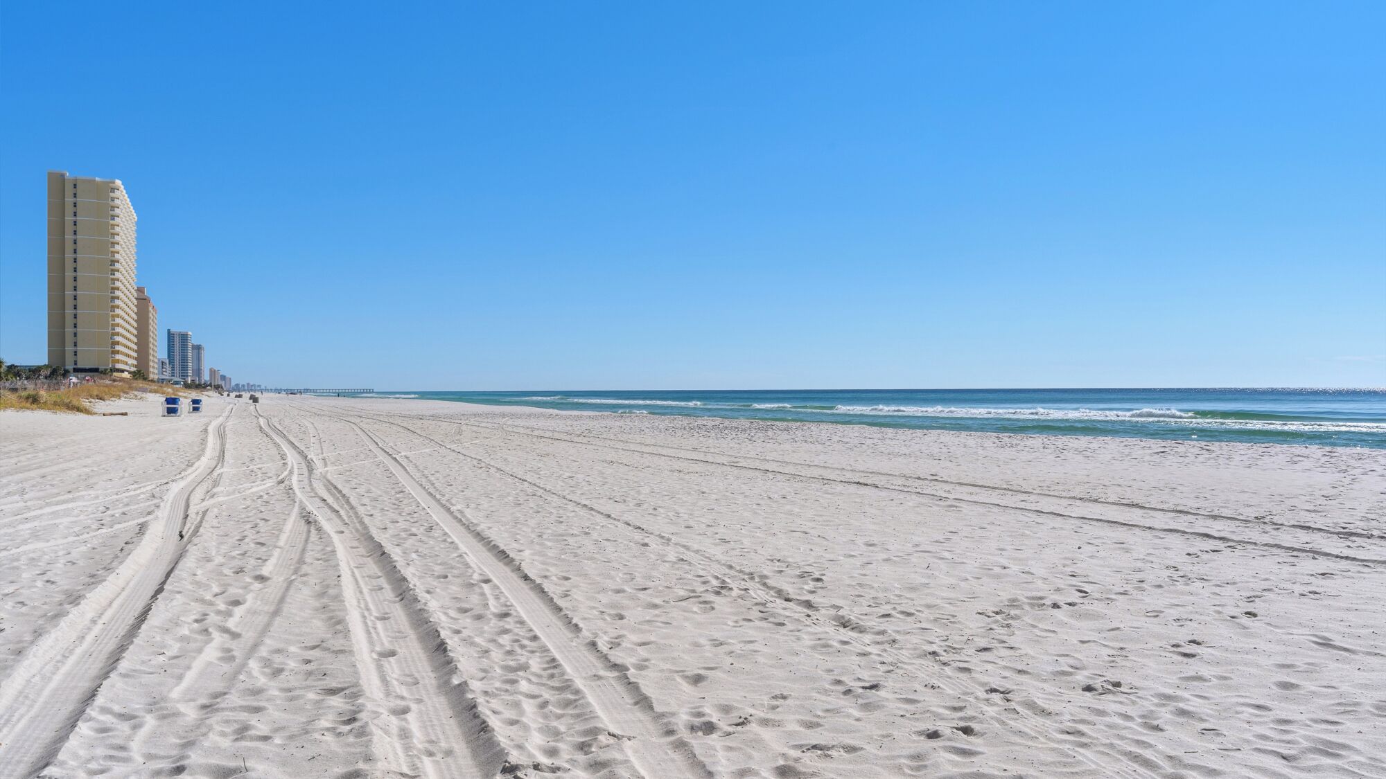 A wide sandy beach with visible tire tracks, a clear blue sky, and a tall building in the background.