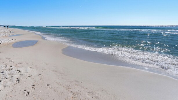 Ein Strand mit Fußspuren, die zum Wasser führen, ein klarer Himmel und ein weiter Pier im Hintergrund.