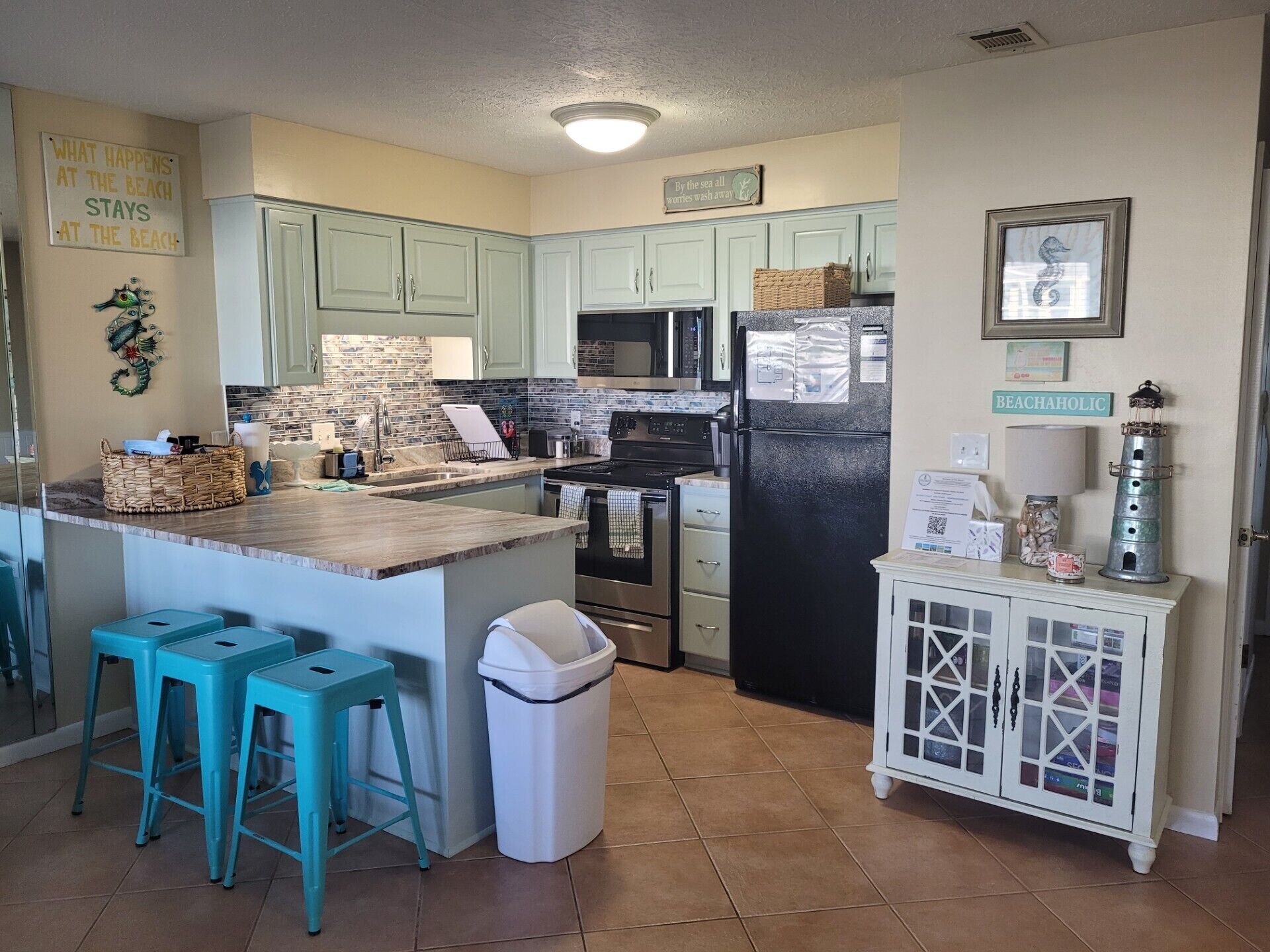 A kitchen with teal cabinets, a black refrigerator, and a small island with stools. There are decorative signs on the walls and a lighthouse decoration on the cabinet.