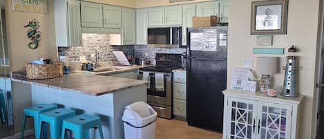 A kitchen with teal cabinets, a black refrigerator, and a small island with stools. There are decorative signs on the walls and a lighthouse decoration on the cabinet.