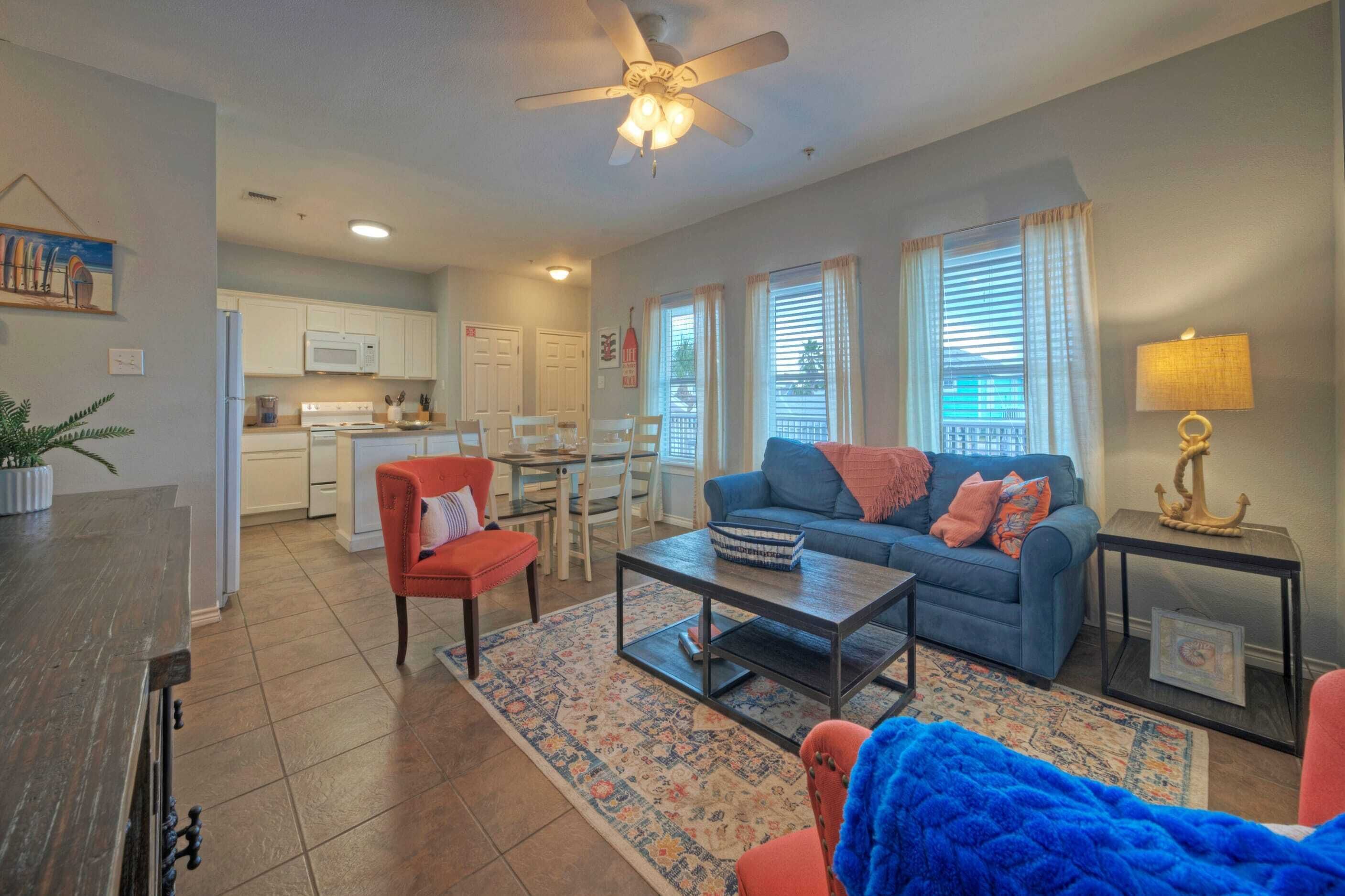 A living room with a sofa, coffee table, and chairs. There is a kitchen in the background with cabinets and a microwave.