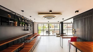 A modern interior with long wooden benches, a central light fixture, and a view through large windows. - Box 150 - LikeHome Hospedagens (João Pessoa)