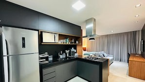 A modern kitchen with a built-in refrigerator, a coffee machine, and a gas stove. There is a small seating area with a sofa and a table. - Box 150 - LikeHome Hospedagens (João Pessoa)