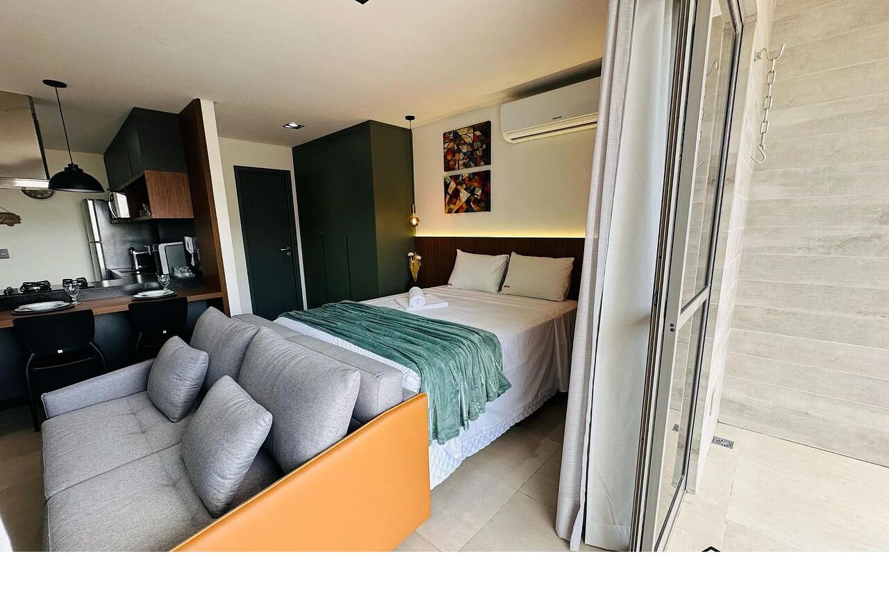 A modern hotel room with a bed, a sofa, a desk with a computer, and a kitchen area.
