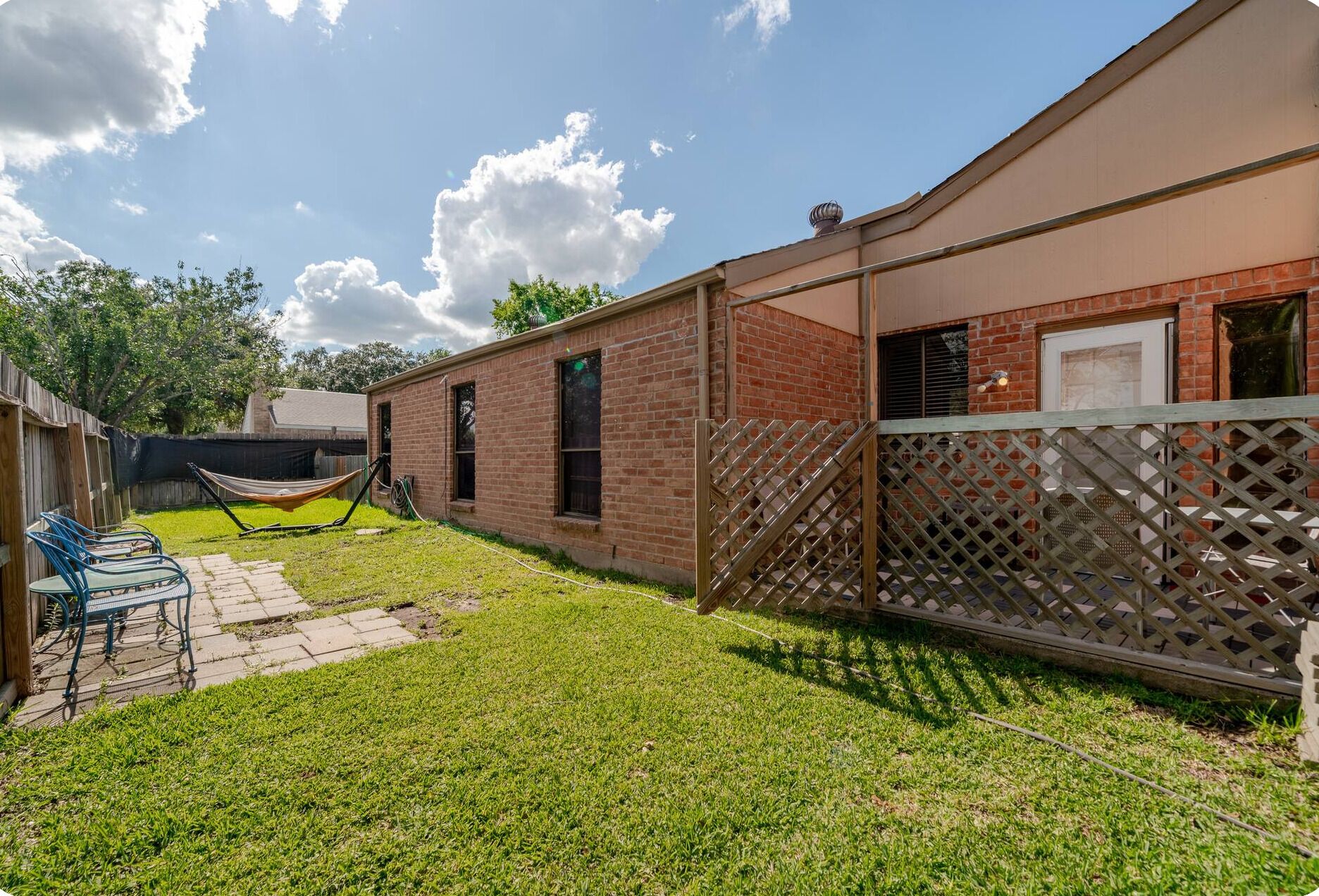 A backyard with a brick house, a hammock, a wooden fence, and a patio with chairs.