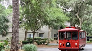 A red trolley moving along a tree-lined street with residential houses in the background. - Walk to Harbour Town in 5 minutes! (Hilton Head Island)