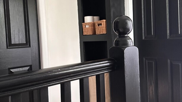 Een zwarte houten deur met een witte omlijsting, een zwarte deurklink en een klein plankje met een mand en opgerolde spullen.