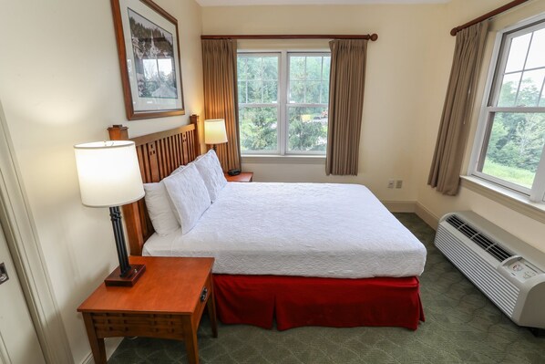 A hotel room with a large bed, a bedside table with a lamp, a window with curtains, and a wall-mounted air conditioner.