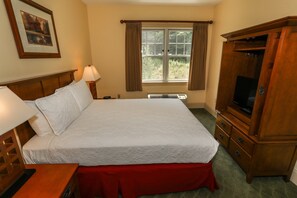 A neatly made bed with a red skirt, a wooden nightstand with a lamp, a television on a wooden cabinet, and a window with curtains.