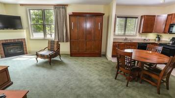 A living room with a fireplace, a dining area with a wooden table and chairs, a kitchen with wooden cabinets and appliances, and a large wooden wardrobe.
