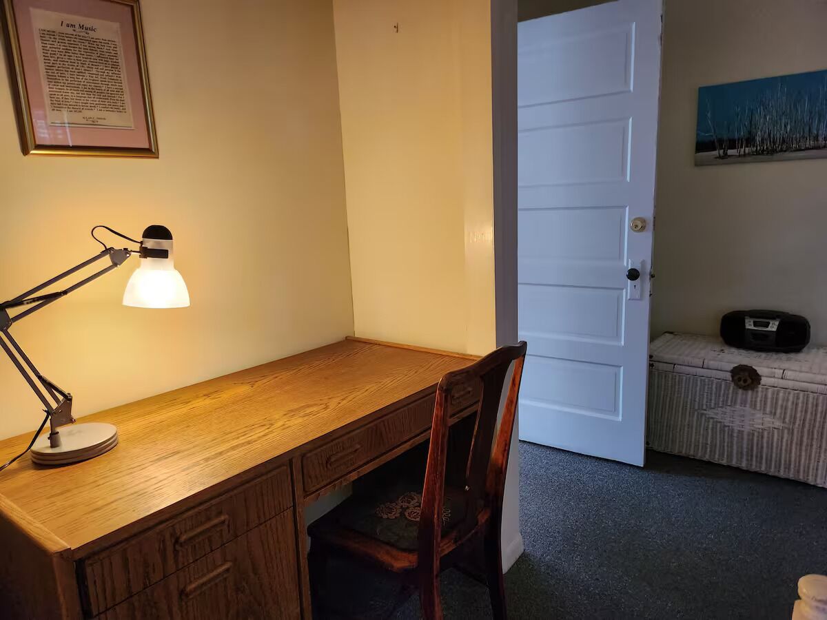 Uma mesa de madeira com luminária, uma cadeira, uma porta branca e um pequeno armário com um rádio.