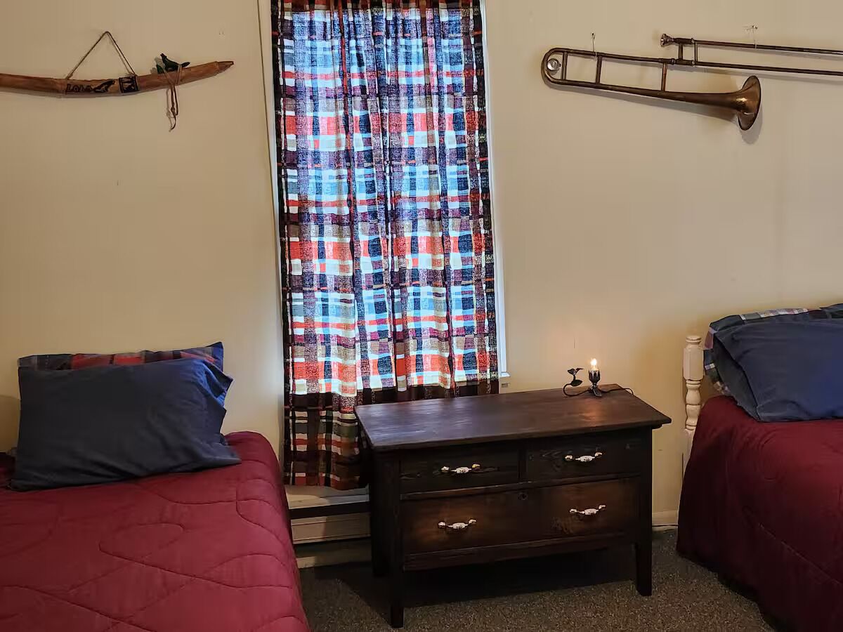 Une chambre avec deux lits, une commode en bois, une fenêtre ornée de rideaux à motifs et un instrument de musique en laiton accroché au mur.