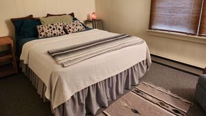 A bedroom with a bed, pillows, a nightstand, a lamp, and a window with blinds.