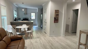 A modern living room with a brown leather sofa, a dining area with a round table and white chairs, and a kitchen with a built-in island and stainless steel appliances.