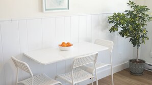 Un comedor con una mesa y sillas blancas, un cuenco de fruta, una obra de arte enmarcada y una planta en maceta.