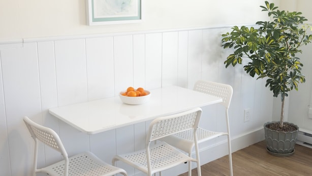 A dining area with a white table and chairs, a bowl of fruit, a framed artwork, and a potted plant.
