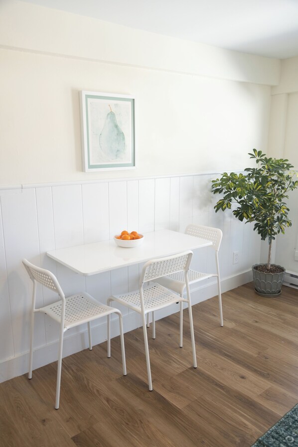 A dining area with a white table and chairs, a bowl of fruit, a framed artwork, and a potted plant. - Peaceful & minutes from the Old Port with private parking (South Portland)