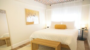 A bedroom with a large bed, a bedside table with a lamp, a bench, a mirror, and a painting on the wall.
