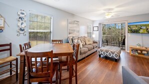 A spacious living room with a wooden dining table and chairs, a sofa with patterned cushions, and a large window with blinds.