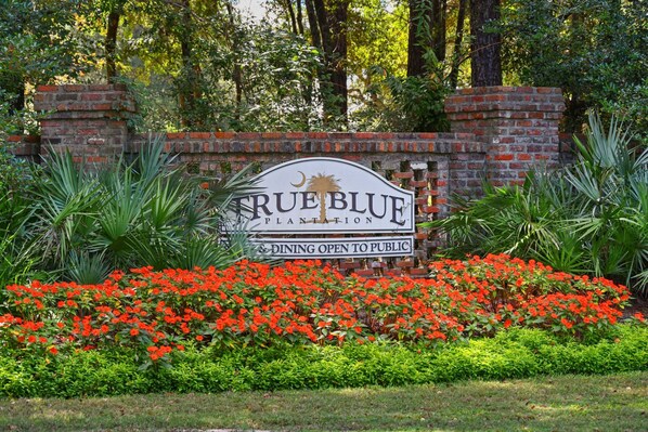 Letrero de True Blue Plantation con un muro de ladrillo y flores rojas en primer plano.