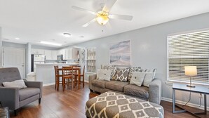 A modern living room with a gray sofa, ottoman, and armchair, a dining area with wooden chairs, and a kitchen in the background.