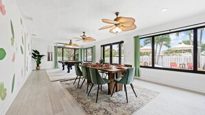 Un vaste espace dédié à la salle à manger, avec une grande table, de nombreuses chaises, des ventilateurs de plafond et de grandes fenêtres offrant une vue sur l’extérieur.