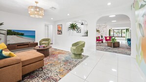 Un vaste espace de vie comprenant un grand téléviseur à écran plat, un fauteuil vert, un tapis à motifs et un luminaire suspendu au plafond.