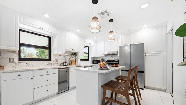 Une cuisine moderne avec des meubles blancs, un grand îlot central, des appareils en acier inoxydable et des luminaires suspendus.