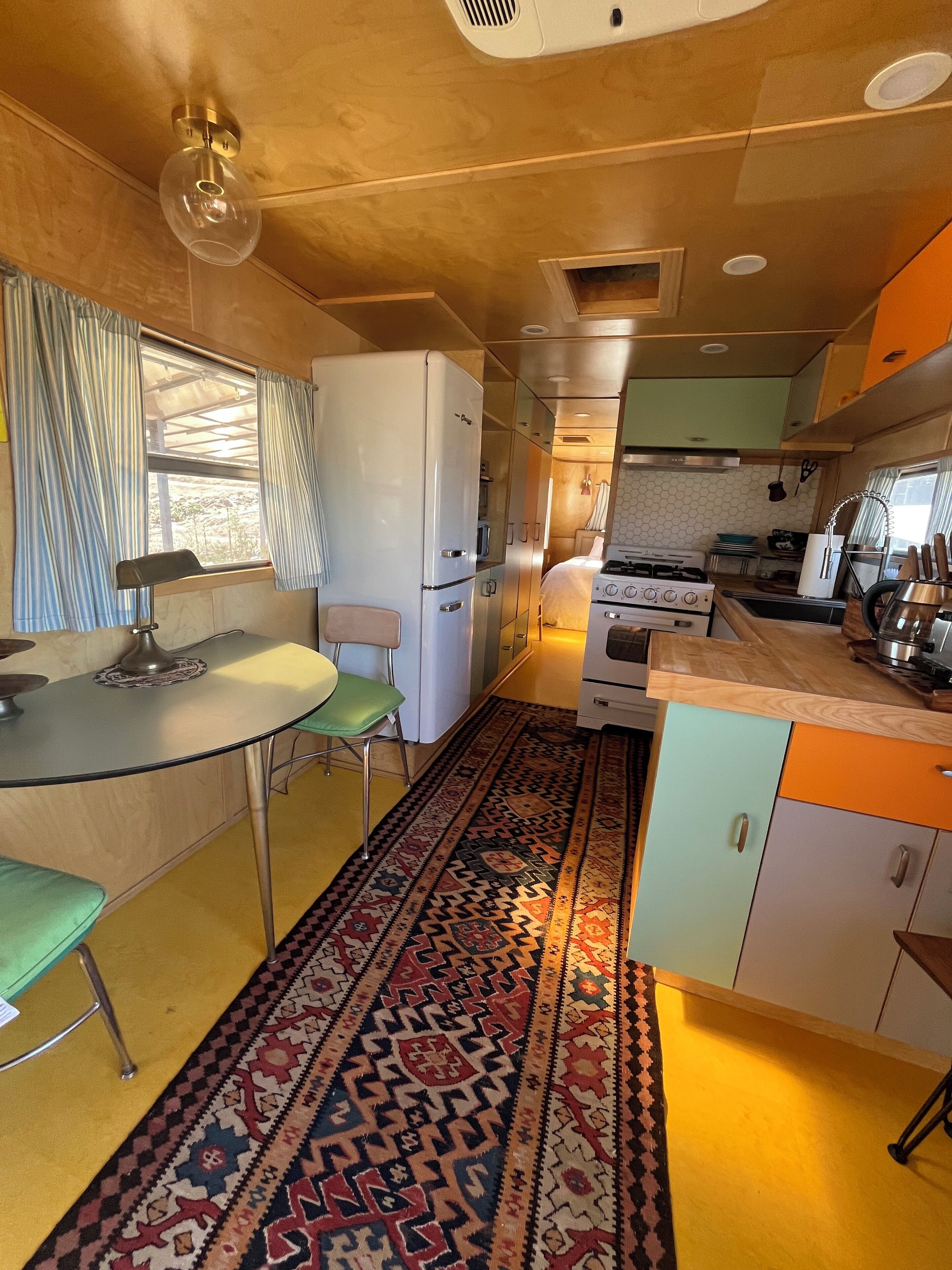 Cocina compacta con refrigerador, estufa y pileta. Se observa una mesa de comedor con sillas y un tapete estampado.
