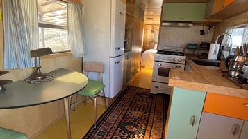 Cocina compacta con refrigerador, estufa y pileta. Se observa una mesa de comedor con sillas y un tapete estampado.