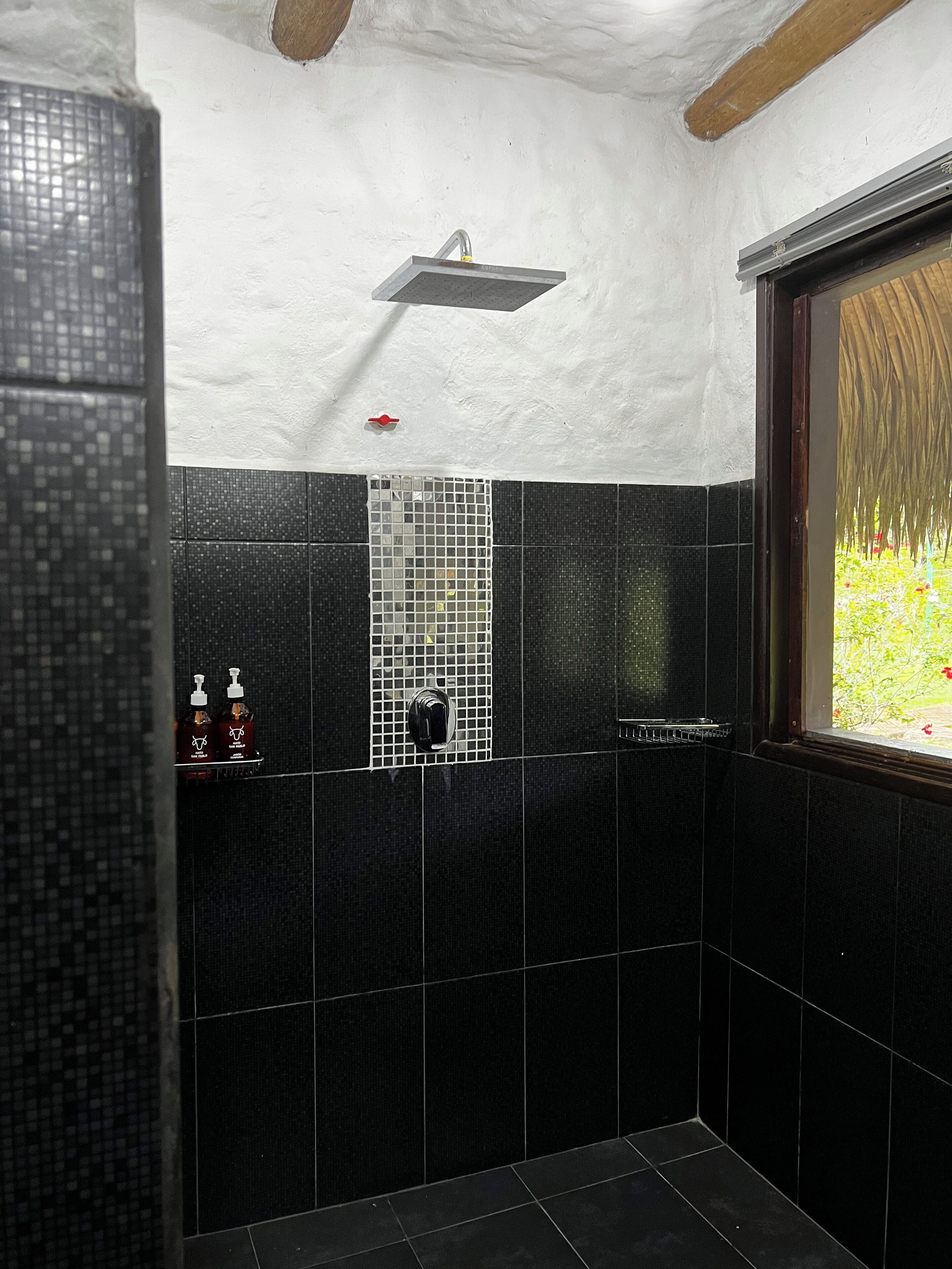 Un baño con azulejos negros, ducha y ventana con vistas a zonas verdes.