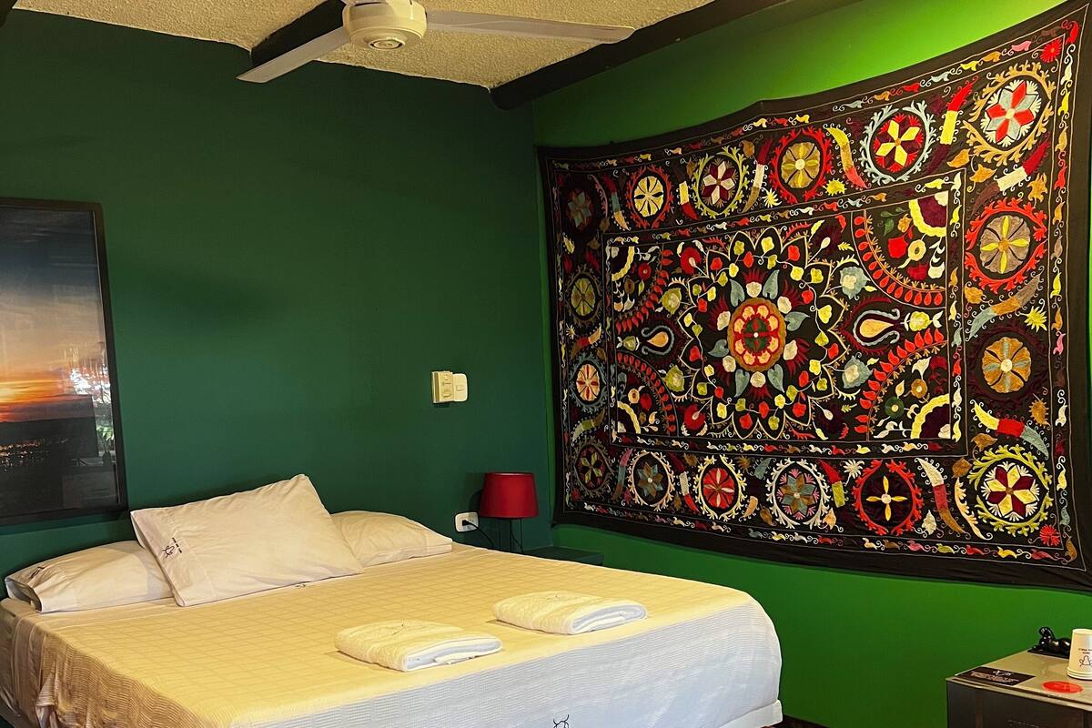 A neatly made bed with white linens, a ceiling fan, a green wall, and a large tapestry.